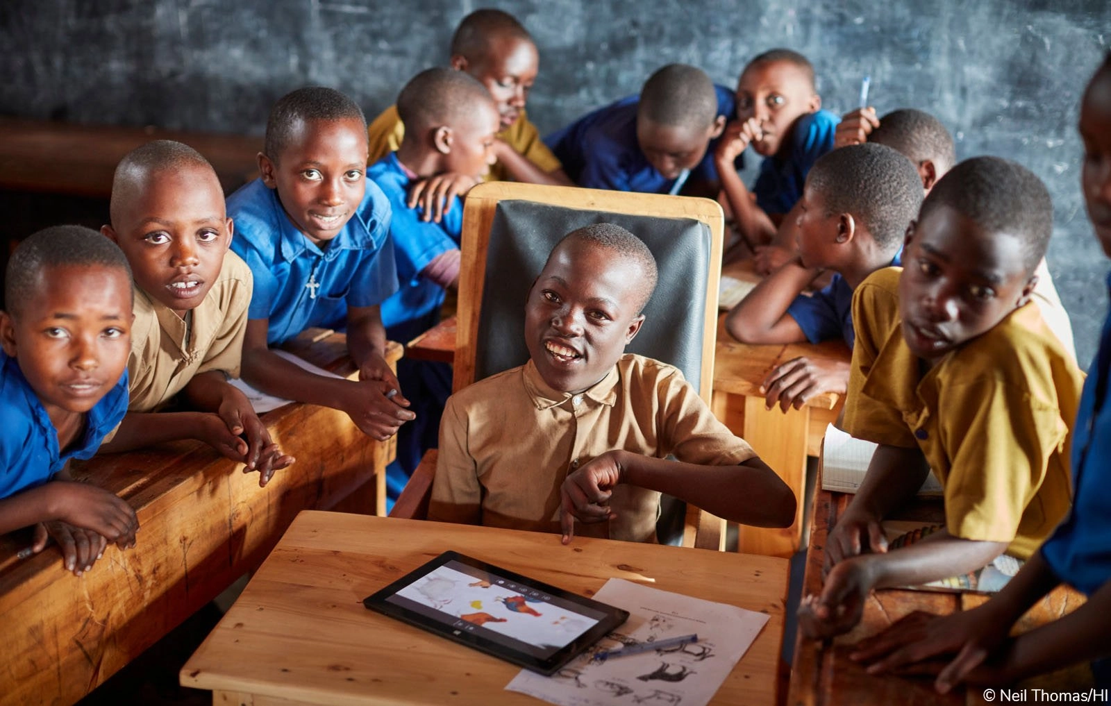 A group of approximately ten young African school-aged boys are gathered around a wooden desk in a dimly lit classroom. A boy seated in a chair in the center is smiling and looking directly at the camera, pointing at a digital tablet placed on the desk in front of him, which shows a colorful application or image. The other boys are leaning in, looking intently at the tablet or the central boy.