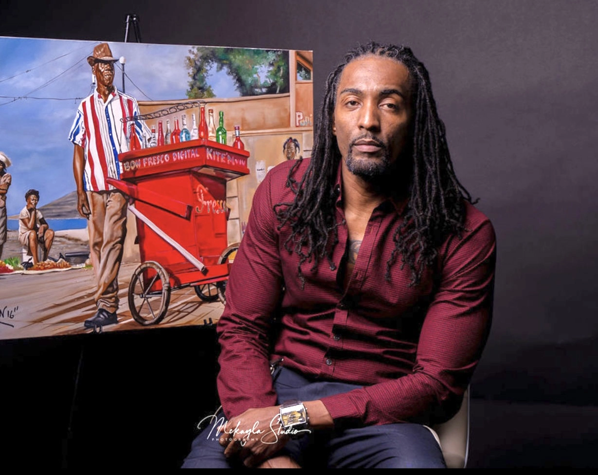 A photograph of a Black man with long dreadlocks and a red long-sleeved shirt, sitting in front of one of his vibrant paintings. The painting features a man in a straw hat pushing a red cart labeled "BON FRESCO DIGITAL" and carrying bottles of beverages on a bright, busy street scene. The photograph is a professional studio portrait.