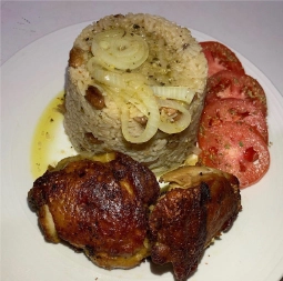 A plate of Cuban-style food featuring a tall, cylindrical mound of seasoned rice, likely arroz congrí or similar, topped with sautéed onion rings and a drizzle of oil. To the right of the rice are three slices of fresh, red tomato. At the bottom of the plate are two crispy, dark-brown pieces of roasted or fried chicken, possibly chicken thighs.