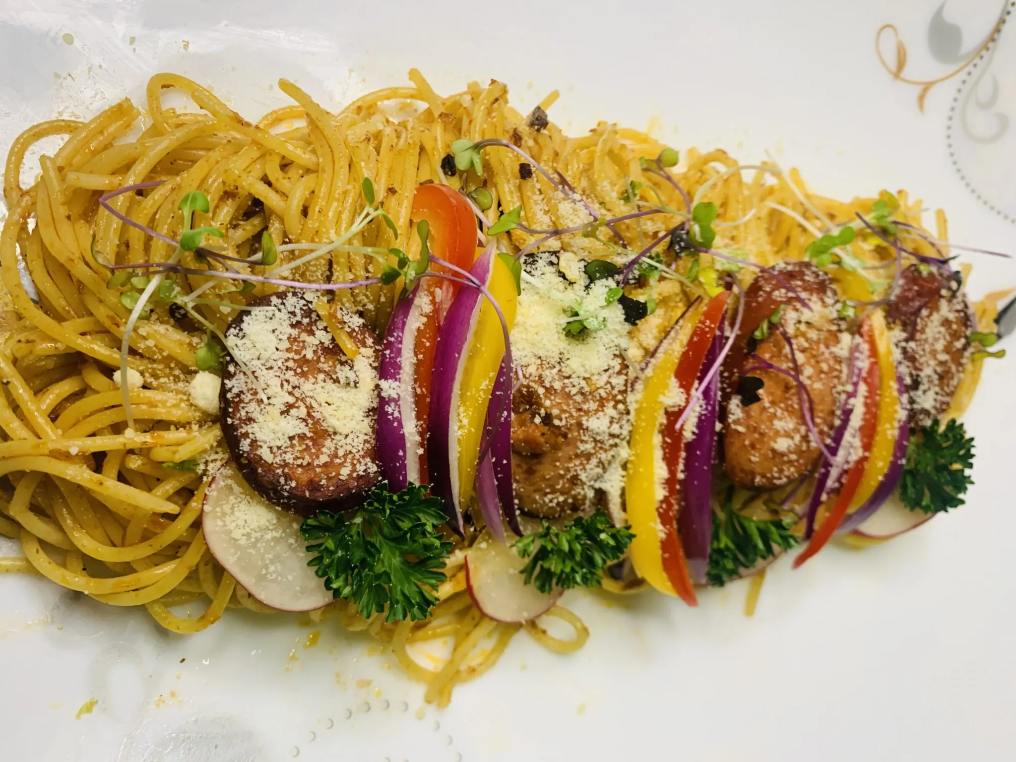 An overhead, close-up shot of a gourmet spaghetti dish served on a long white platter. The pasta is lightly dressed and topped with thick slices of grilled sausage, which are separated by colorful vertical slices of bell peppers (red, yellow) and red onion. The dish is garnished with microgreens, grated Parmesan cheese, fresh parsley, and thin slices of radish.