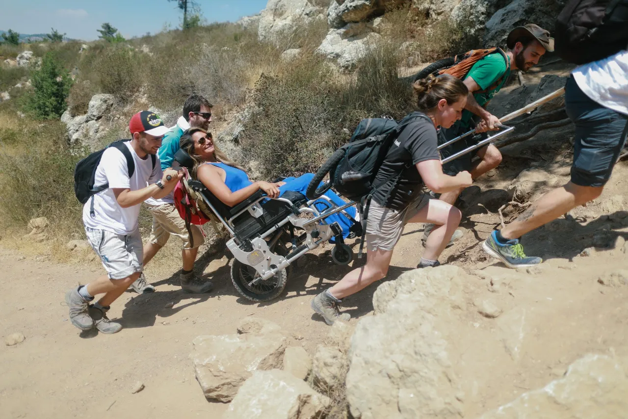 A group of six hikers ascends a steep, rocky, sunlit dirt path. Four members of the group are pushing and pulling a specialized, all-terrain wheelchair occupied by a woman in a blue top who is smiling and wearing sunglasses. The path is rocky and surrounded by dry shrubs and trees.