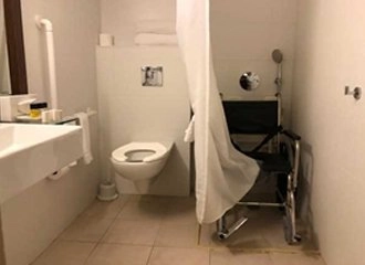 An image of an accessible bathroom. The room features a wall-mounted toilet, a sink with an accessible clearance underneath, and white grab bars. A rolling shower chair is visible in the shower area, which is partially separated by a white shower curtain.