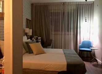 A dimly lit hotel room furnished with two twin beds pushed together, made with white and teal linens and topped with several pillows. The beds are facing large, sheer curtains covering a window. A small, round side table and a bright blue accent chair are visible on the right.