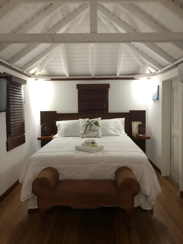 An interior view of a bright hotel bedroom featuring a queen-sized bed with white linens centered under a high, white, pitched ceiling. A dark brown headboard and two small nightstands flank the bed. A brown, velvet bench sits at the foot of the bed, and dark wooden blinds cover the windows. A decorative pillow on the bed reads "Pasture Piece."