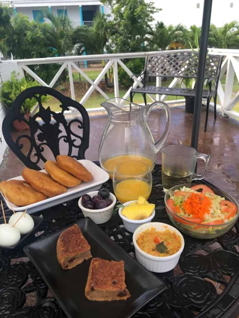 A close-up view of a large breakfast spread laid out on a black, ornate metal outdoor table on a patio. The items include a platter of golden-brown fried dough (possibly Johnny Cakes or bakes), two slices of fruit or spice cake on a black plate, a bowl of seasoned vegetables or hash, a clear glass pitcher and cup of orange juice, a cup of clear tea, a bowl of grapes, sliced pineapple, two boiled eggs on toothpicks, and a large bowl of fresh garden salad. Palm trees and a fence are visible in the background.