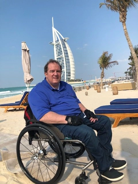 A middle-aged man in a blue polo shirt and jeans is seated in a manual wheelchair on a sandy beach. He is looking forward and wearing black gloves. In the background, the iconic sail-shaped Burj Al Arab hotel in Dubai stands tall against a clear blue sky. Palm trees and beach loungers are visible nearby.