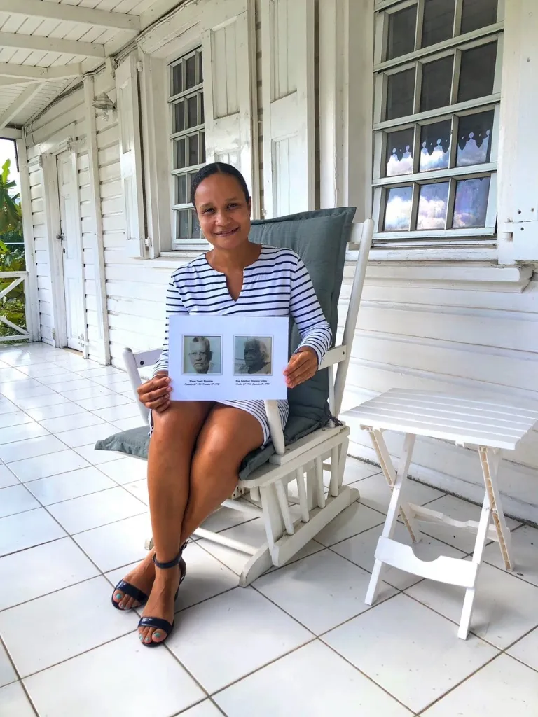 A woman in a striped short dress and dark sandals sits on a white rocking chair on a white-tiled porch, holding a small white sign with two framed black-and-white portraits and text, with her eyes directed toward the camera. White wooden walls and shuttered windows are visible behind her.