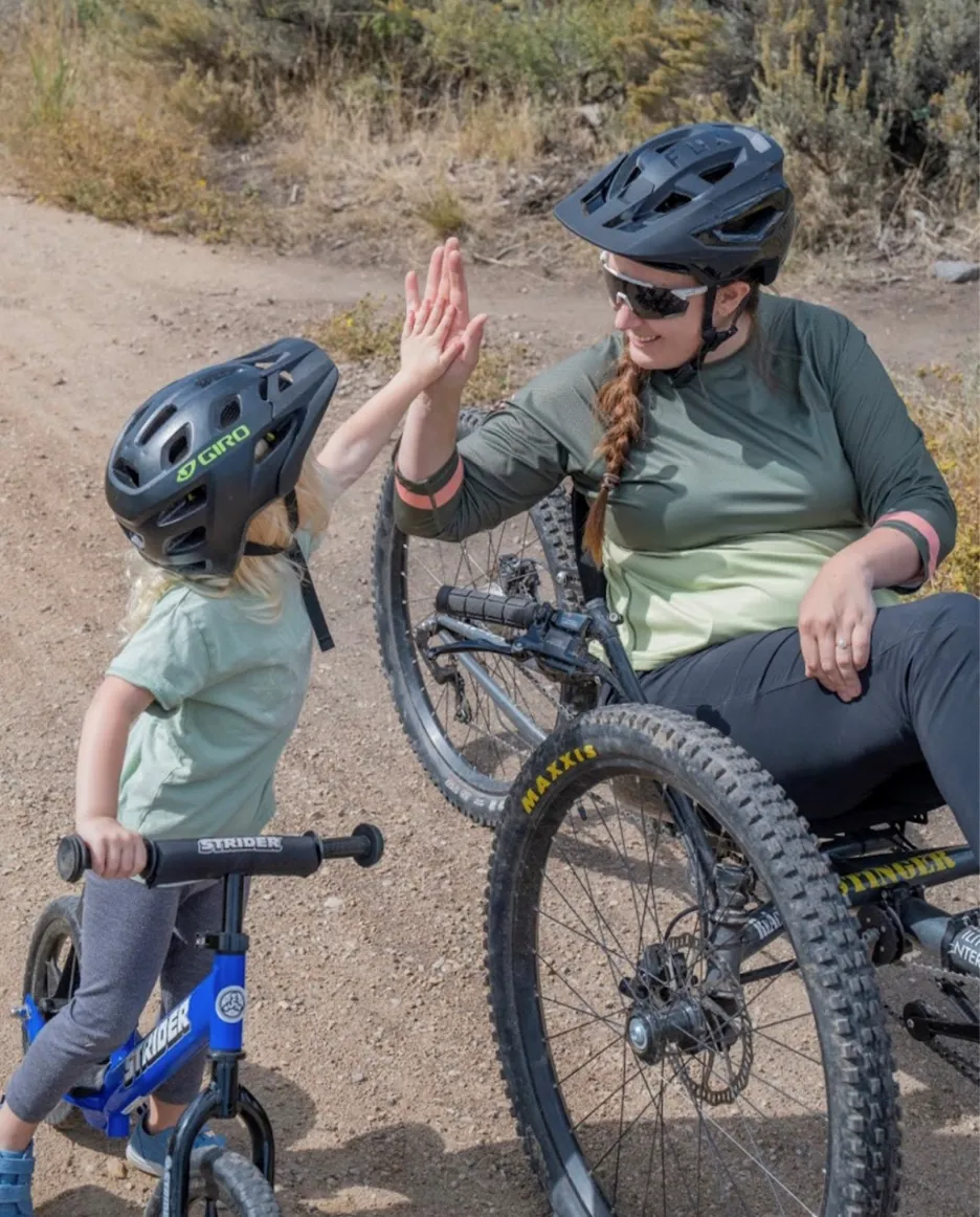On a dirt and gravel path, a woman in a 3-wheeled wheelchair high-fives a young girl on a small blue bike.