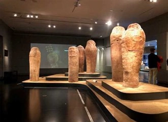 An interior view of a museum exhibit showcasing several tall, ancient anthropoid clay sarcophagi (funerary coffins). The sarcophagi are standing on elevated, curved wooden platforms in a dimly lit room with a dark floor, and a projection screen is visible on the