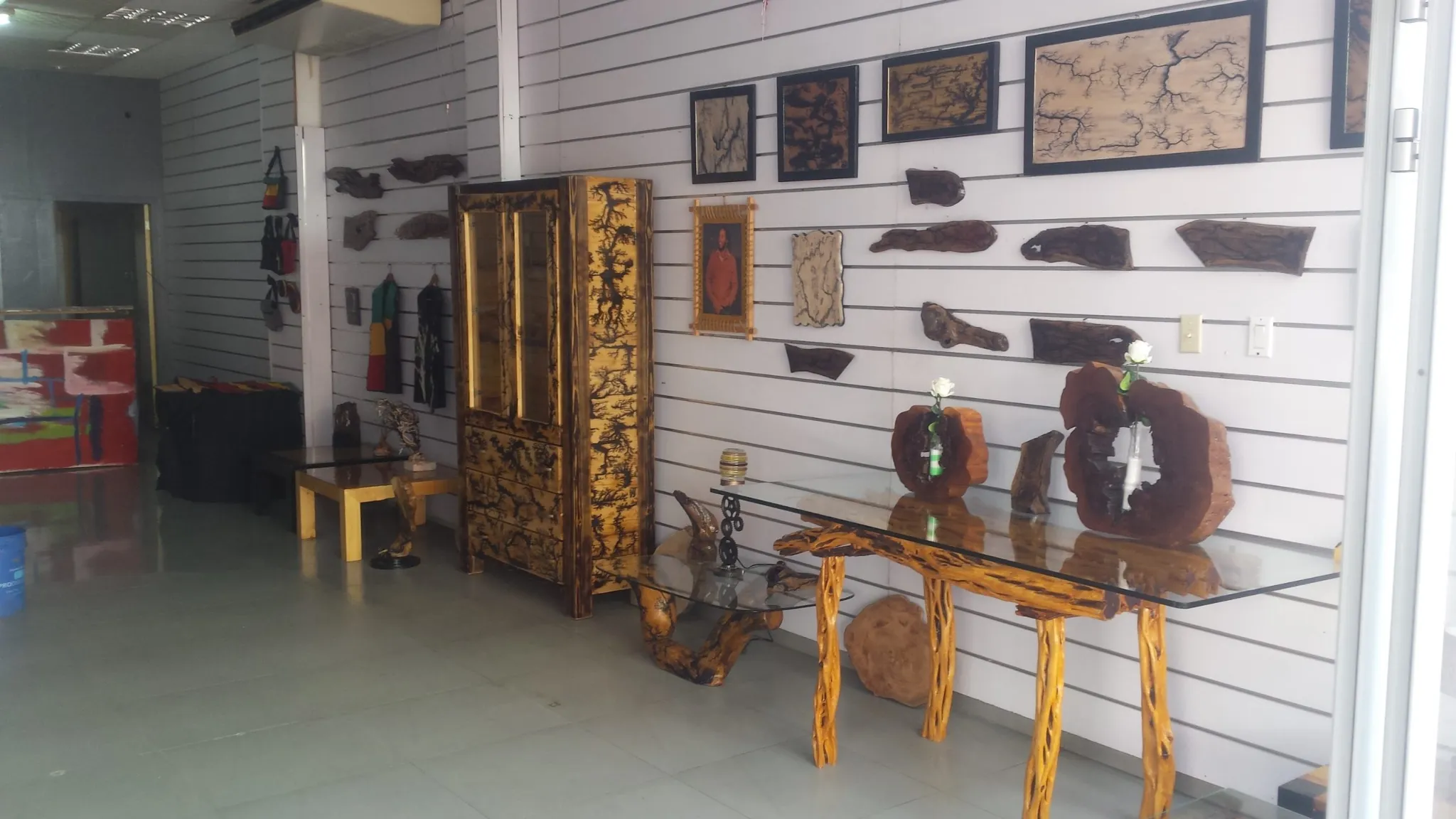 The interior of a retail space or gallery showcasing custom furniture and artwork. A tall wooden cabinet and two console tables with glass tops are visible, all crafted from natural wood with distinctive Lichtenberg figures (electric burns). Various pieces of wood art and wall hangings are displayed on the white slatted wall.