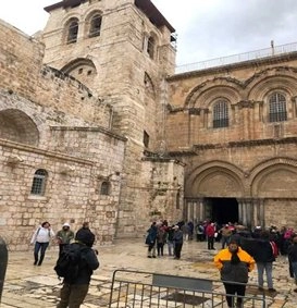 The exterior entrance facade of the Church of the Holy Sepulchre in Jerusalem, featuring a large stone belfry and Romanesque arches over the entrance doors. A crowd of pilgrims and tourists in various jackets is gathered in the stone courtyard in front of the entrance.