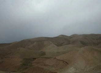 A wide, expansive view of undulating, tan and brown desert hills under a heavy, overcast, gray sky, suggesting a landscape in the Judean or Negev Desert.