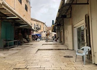 A narrow, stone-paved street in an outdoor marketplace, likely in the Old City of Jerusalem. The street is lined with shops under awnings, many with closed or partially closed metal shutters. A single light gray plastic chair sits on the right side of the street. The sky is cloudy.