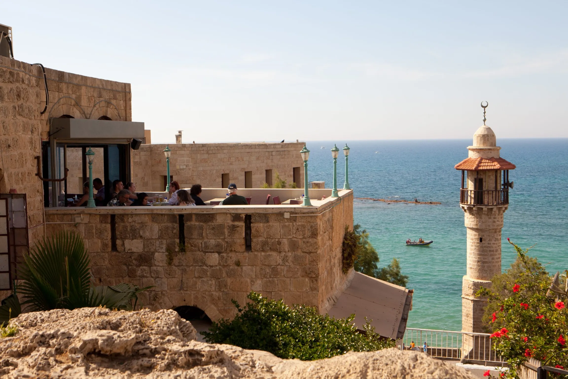 A sunny outdoor scene of a restaurant patio built into the old stone walls overlooking the Mediterranean Sea. A group of people are seated at a table on the terrace. To the right, a distinctive stone minaret with a small balcony and a crescent moon on top stands next to the water, with a small boat visible in the blue sea.