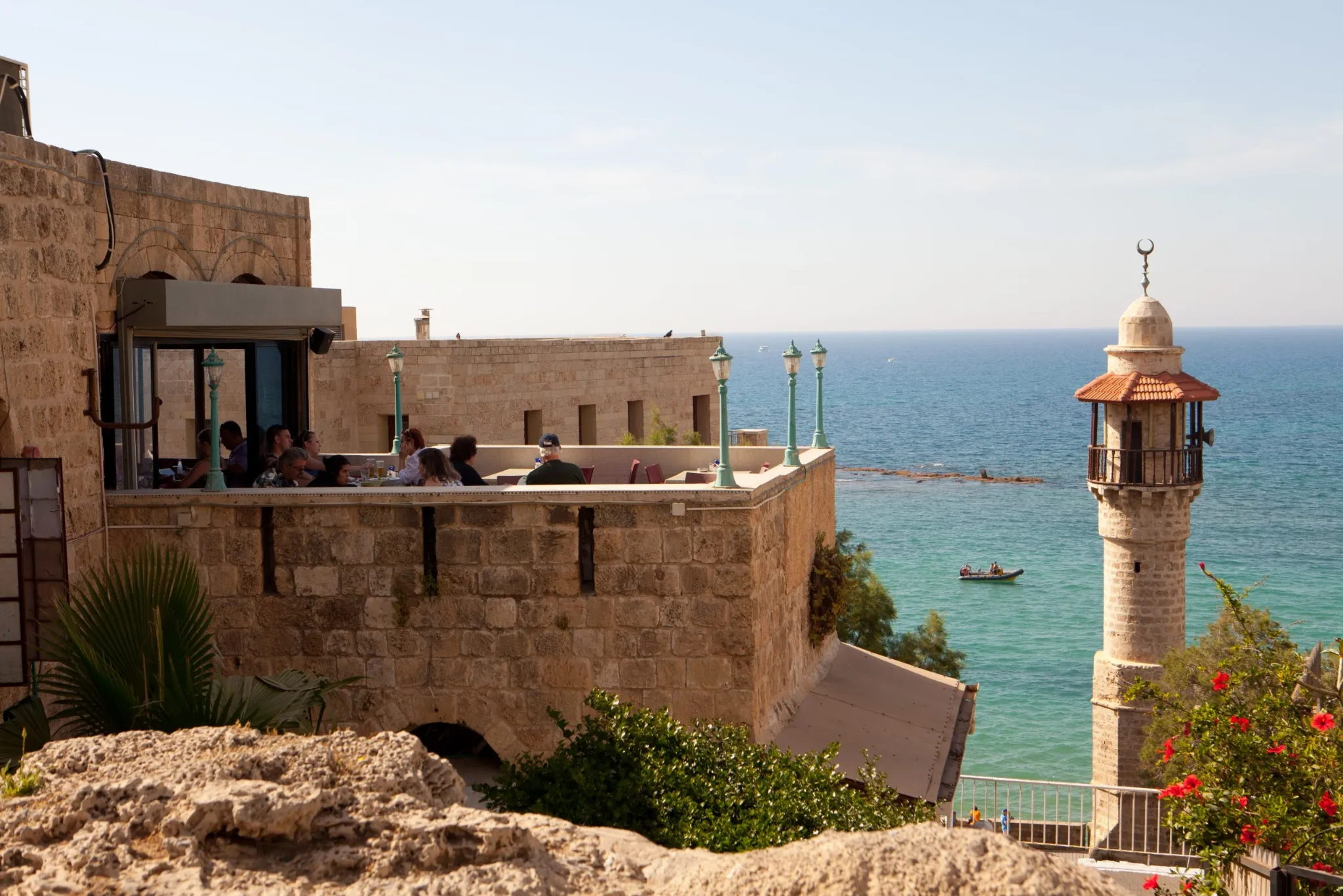 A sunny outdoor scene of a restaurant patio built into old stone fortifications overlooking the blue sea. A group of people are seated on the terrace. To the right, a distinctive stone minaret with a small balcony and a crescent moon stands next to the water, with a small boat visible in the sea.