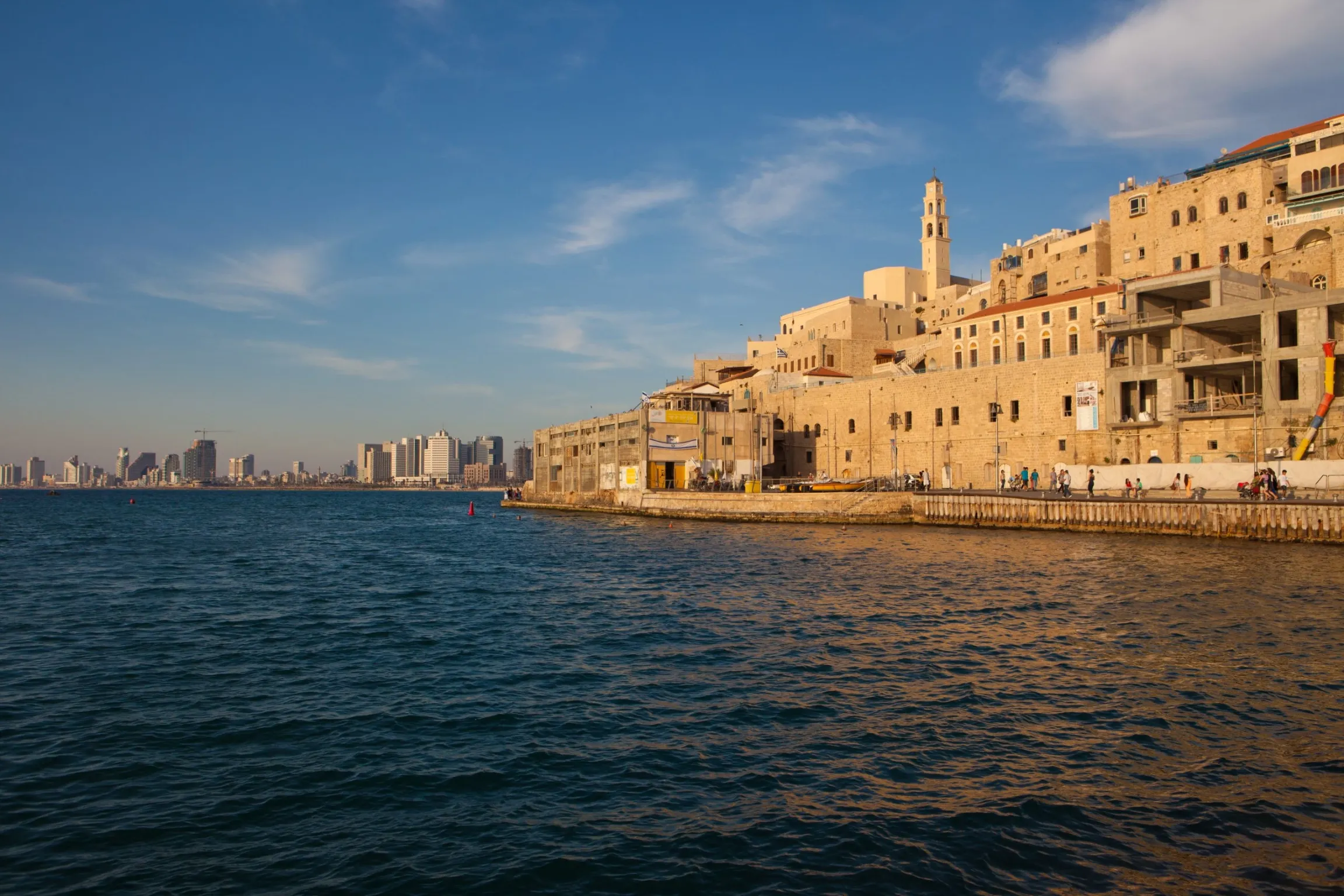 A view of the historic sea wall and ancient buildings of Jaffa (or Old Tel Aviv) from the water, under a blue sky. The ancient stone buildings, including a prominent minaret, line the coast, with the modern Tel Aviv skyline visible in the distance across the bay.