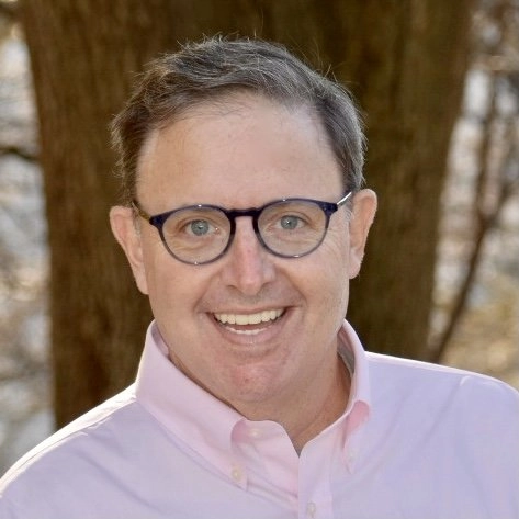 A headshot of a middle-aged man with short brown hair, wearing a light pink collared shirt and blue-rimmed glasses. He is smiling warmly at the camera, standing outdoors with the blurred background of a tree trunk and foliage.