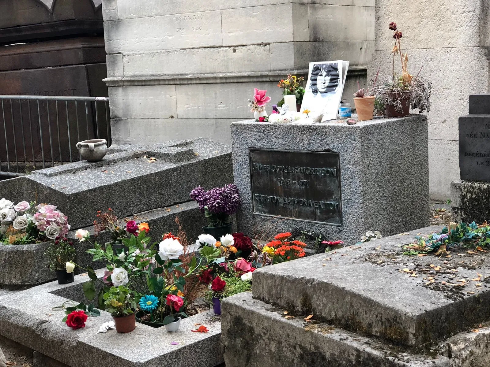 A close-up photo of the grave of Jim Morrison at Père Lachaise Cemetery in Paris. The central grave marker is a simple, granite cuboid with a bronze plaque inscribed with the name JAMES DOUGLAS MORRISON / 1943-1971 and the Greek phrase KATA TON DAIMONA EAYTOY. The grave is surrounded by flowers in pots and scattered petals, and a framed black and white photo of a young Jim Morrison rests on the stone marker. Other older, gray stone tombs are visible on either side and behind it.