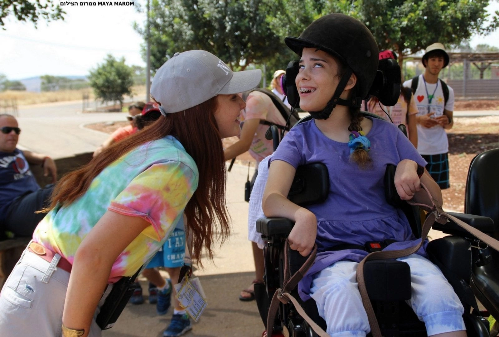A candid outdoor photo showing a smiling young woman with long brown hair and a gray baseball cap leaning down to speak to a girl seated in a wheelchair. The girl in the wheelchair is wearing a black helmet and a purple shirt and is smiling and looking up at the woman. They are both outside on a sunny day in a recreational area with trees and other people visible in the background.