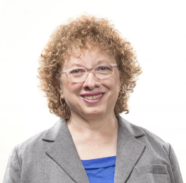 A close-up, professional headshot of an older woman with short, curly reddish-blonde hair wearing glasses, a bright blue top, and a grey blazer, smiling warmly against a white background.