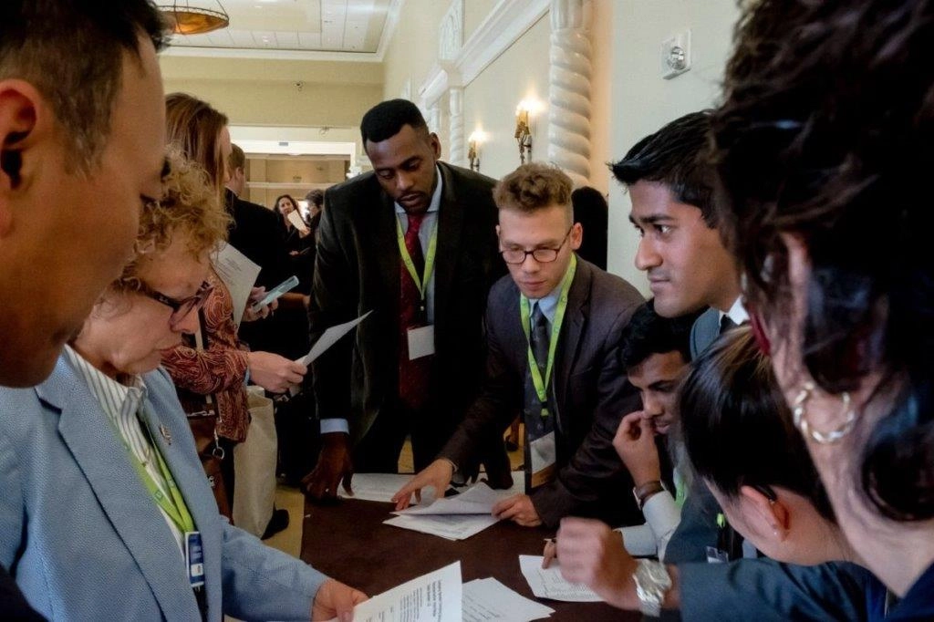 A group of approximately ten professionals, mostly men in business attire, huddle around a table covered in papers and documents, intently reviewing them in what appears to be a formal conference or event setting. Two men in the center are pointing at the documents.