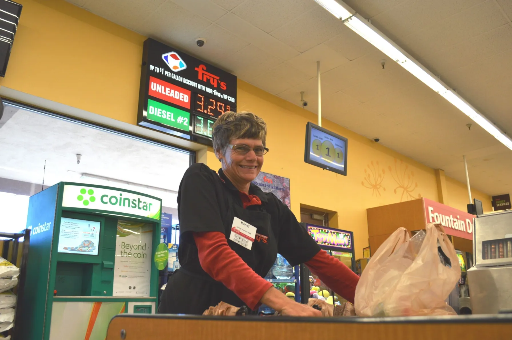 A friendly, middle-aged female grocery store employee with short hair and glasses, wearing a black uniform shirt over a red long-sleeve shirt, smiles while standing behind a checkout counter bagging groceries. In the background, a large digital sign displays fuel prices for 'Unleaded' and 'Diesel #2' with a "Fry's" logo, and a green Coinstar machine is visible on the left.