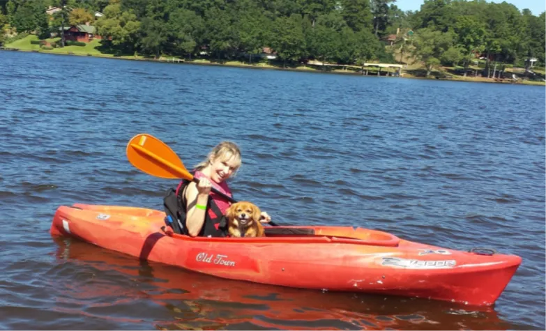 Under a sunny blue sky, a woman paddles a red kayak on a lake. She smiles from the cockpit, a small dog at her side.