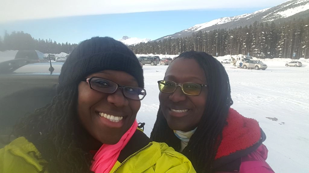 A close-up selfie of two Black women smiling and standing outdoors in a bright, snowy landscape. The woman on the left wears a black knit hat and glasses, a neon yellow jacket, and a bright pink scarf. The woman on the right wears yellow-tinted glasses and a pink/red winter jacket. In the background, there is a parking lot filled with cars, snow-covered ground, and a pine forest leading up to snow-capped mountains under a clear blue sky.