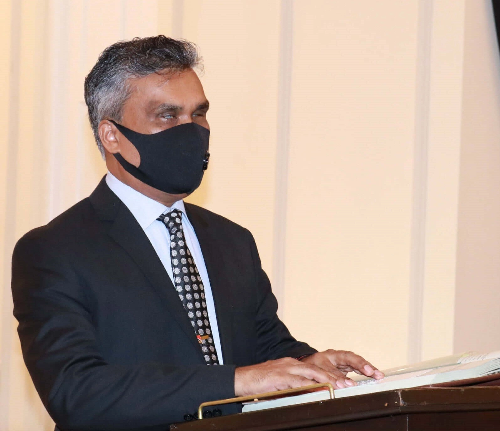 A middle-aged man with salt-and-pepper hair, wearing a dark suit, a patterned tie, and a black face mask, standing behind a wooden lectern or table with his hands resting on an open book or document. He is looking off to the side, and the background is a simple, light-colored interior, likely a formal setting.