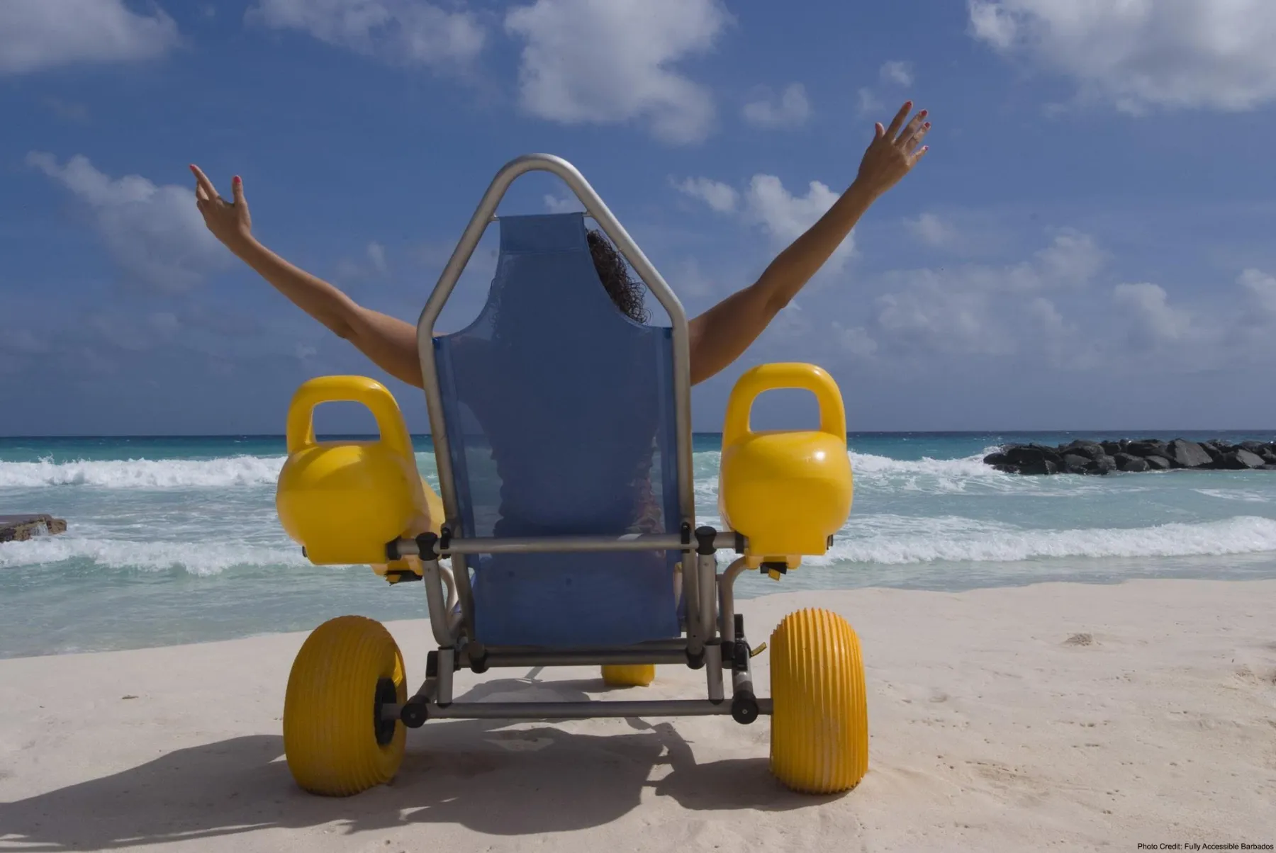 A person sitting in a specialized, blue beach wheelchair with large, yellow, balloon-like wheels, facing away from the camera on a white sand beach. The person has their arms raised overhead in a gesture of triumph or excitement, looking out at the turquoise ocean waves. The sky is bright blue with scattered white clouds.