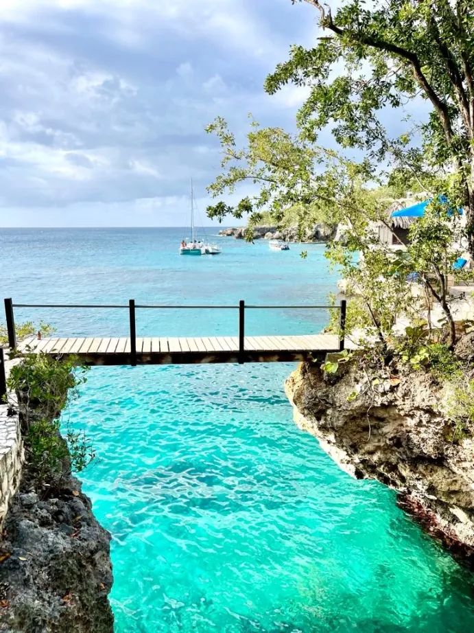A picturesque scene of turquoise water on a cloudy day, framed by rocky cliffs and lush green trees. A small wooden footbridge with black railings crosses a narrow gap between the cliffs over the brightly colored ocean channel. In the distance, a white sailboat is anchored in the bay.