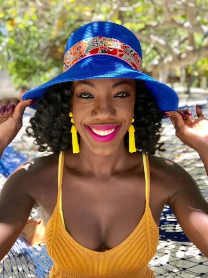 On a sun-dappled patio, a smiling woman grasps her vibrant blue hat. She wears hot pink lipstick, bright yellow earrings and knitted top.