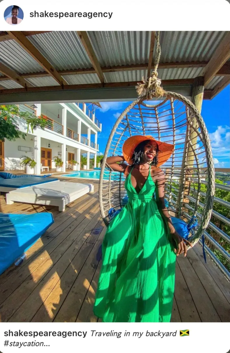 An image posted by the user "shakespeareagency" showing a smiling woman of color wearing a bright green maxi dress and a wide-brimmed orange hat, sitting in a woven egg-shaped hanging chair on a wooden deck. In the background is a modern white two-story building with a poolside patio and a swimming pool, visible under a sunny blue sky. The caption reads "Traveling in my backyard" followed by a Jamaican flag emoji and the hashtag #staycation.