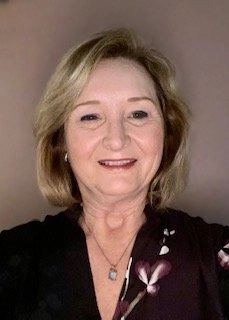 A head-and-shoulders portrait of a middle-aged woman with short, blonde hair, smiling softly at the camera. She is wearing a dark top with a purple and white floral pattern and a small pendant necklace. The background is a plain, light brown color.