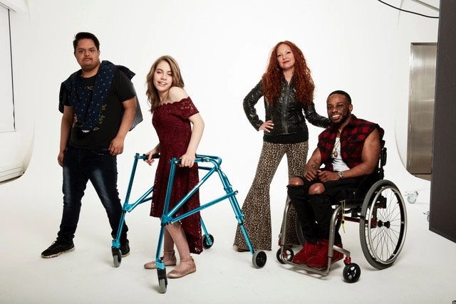 A studio portrait of four individuals posing on a white background. From left to right: a person stands wearing dark clothes and a patterned shawl; a smiling teenage girl in a maroon dress stands holding a metallic blue wheeled walker; a woman with long red hair wearing a black leather jacket and leopard-print flared pants stands with her hands on her hips; and a man in a wheelchair wearing a red and black plaid vest over a black shirt and torn jeans sits smiling.