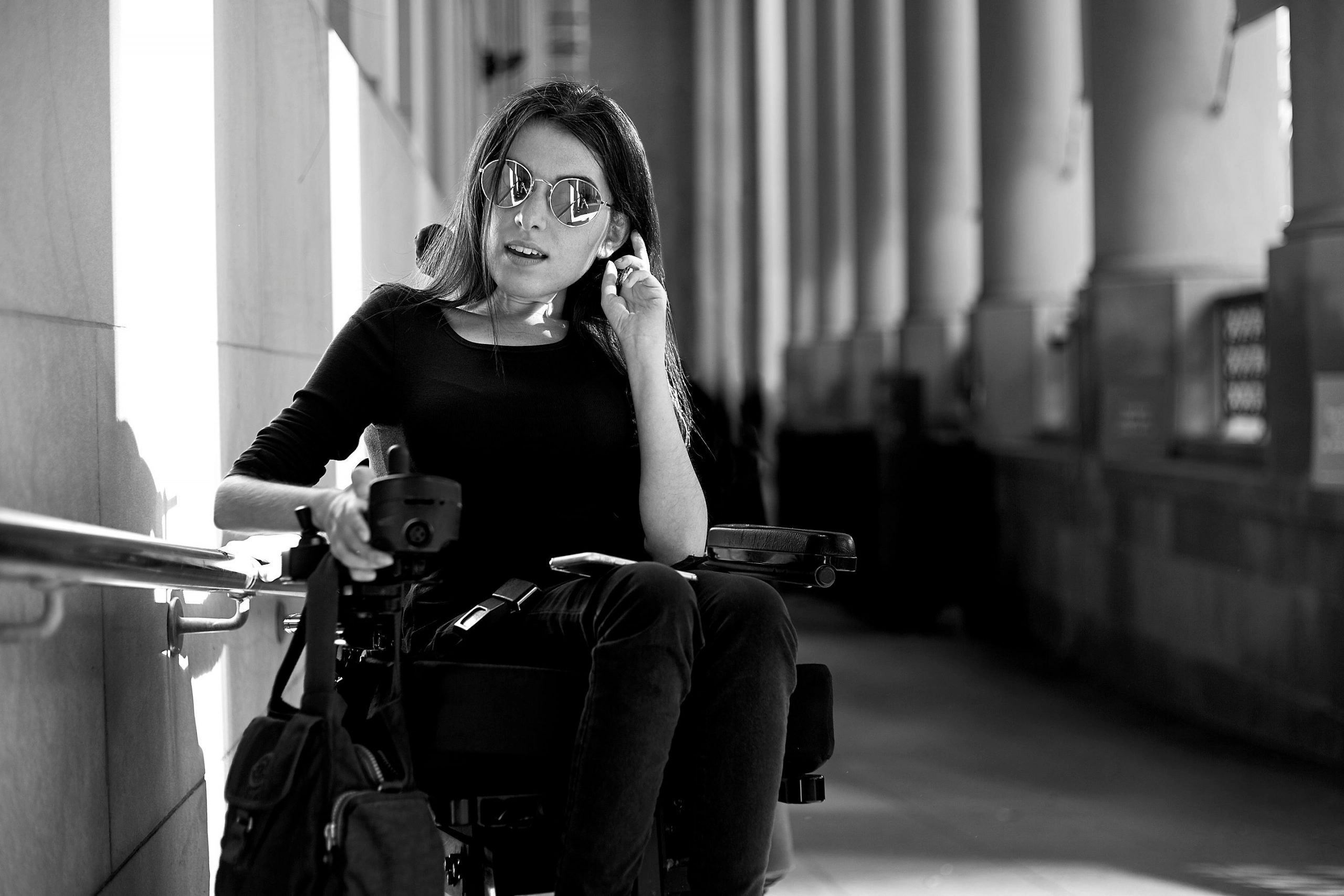 Black and white portrait of a young woman seated in a power wheelchair wearing dark clothing and large round sunglasses. She is outdoors in a columned arcade, touching her ear with one hand.