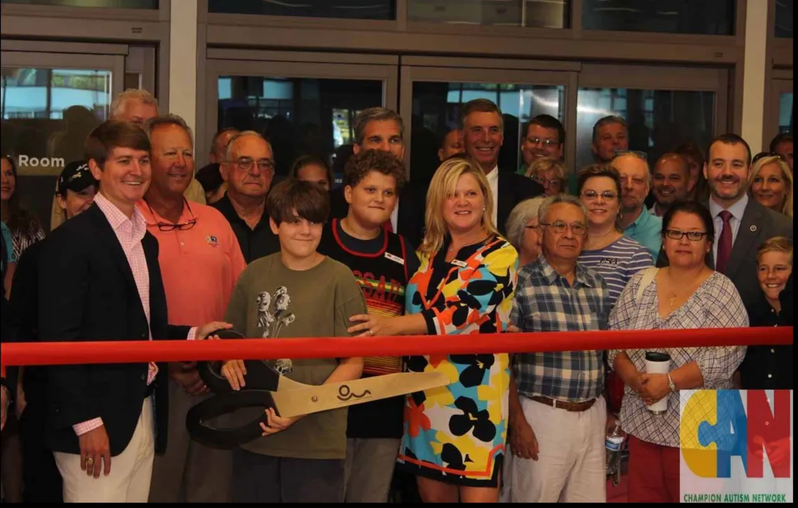 A group of diverse adults and children are gathered inside a building for a ribbon-cutting ceremony. Two young boys stand in the center, one holding oversized novelty scissors ready to cut a thick red ribbon stretched across the foreground. They are surrounded by smiling attendees, including a woman in a brightly colored dress, and several men in suits and casual wear. The atmosphere is celebratory, and a logo for the 'Champion Autism Network' is visible in the bottom right corner.
