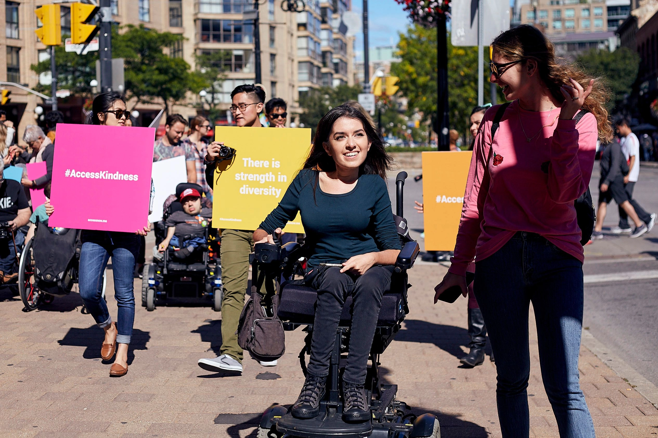 A woman with long dark hair, wearing a dark green top, sits in a motorized wheelchair on a sunny city street. She is smiling and surrounded by a diverse group of people holding bright pink and yellow signs with messages about accessibility and inclusion, such as 'There is strength in diversity.'