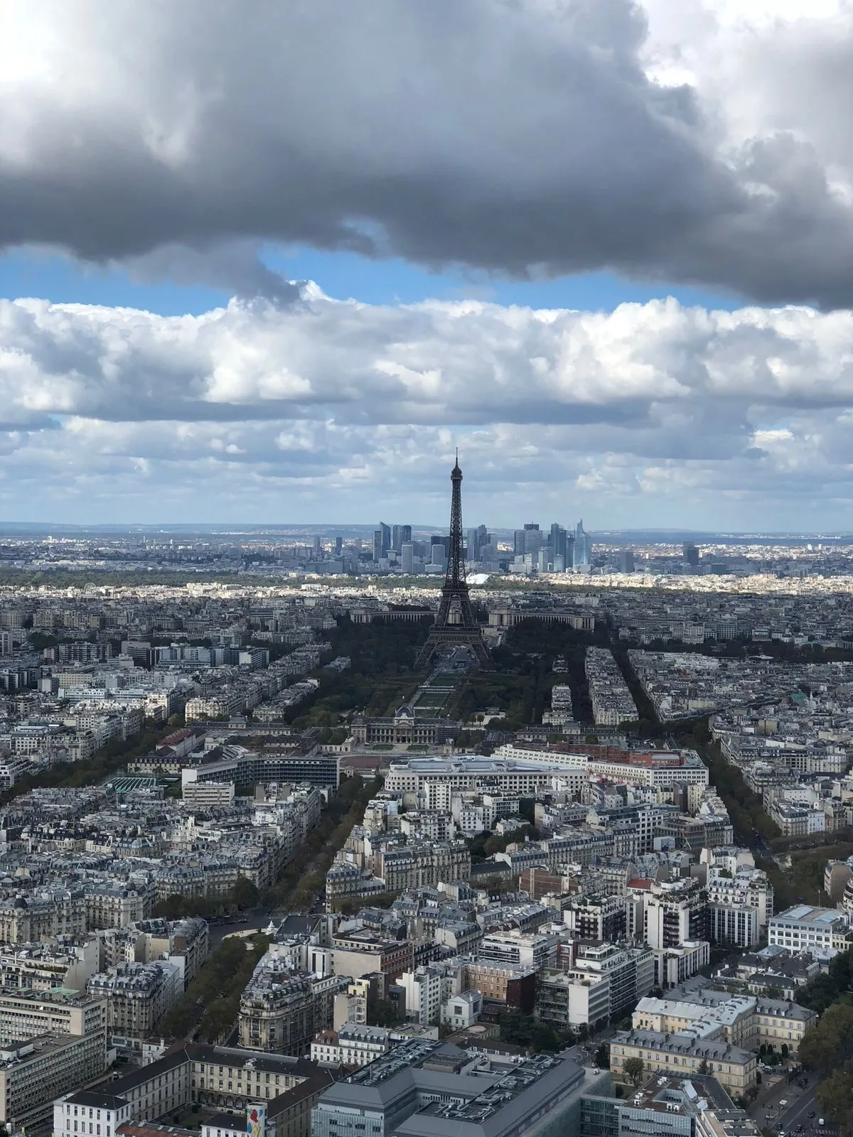 A sweeping, high-angle panoramic view of the Paris cityscape taken from a skyscraper, likely the Montparnasse Tower. The Eiffel Tower stands centrally in the middle distance, surrounded by low-rise gray buildings. Beyond the Eiffel Tower, the modern skyscrapers of the La Défense business district are visible on the horizon. The sky is dramatic with large patches of white and blue clouds.
