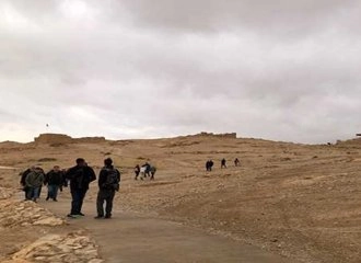 A group of tourists walking on a paved pathway across the vast, arid, brown terrain at the top of Masada, Israel, toward the ancient ruins visible in the distance. The sky is heavily overcast and gray.