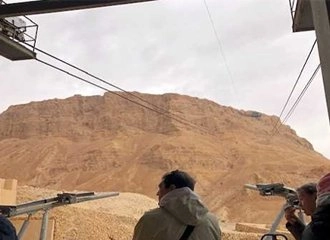 A view from a cable car station looking up at the massive, light brown desert mountain of Masada, Israel. Overhead, the cable car wires stretch toward the peak. The foreground shows the heads and shoulders of a few people waiting, looking up at the mountain under a cloudy sky.