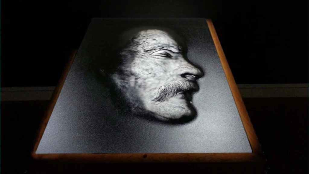 A highly textured, close-up profile portrait of an older man with a pronounced mustache and beard, illuminated by dramatic light from the front, creating a stark black-and-white contrast. The image appears to be a tactile, raised surface designed to be felt.
