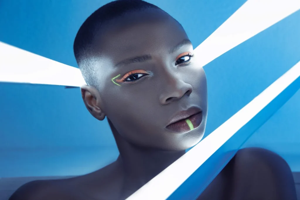 A close-up, futuristically styled portrait of a person with a buzz cut and dark complexion, centered between bright white light beams against a blue background. They are wearing neon orange eyeshadow, a neon green arrow graphic near the eye, and a thin neon green vertical line accent on their lower lip.