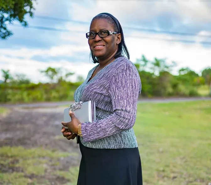 A medium outdoor portrait of a smiling woman wearing black-rimmed glasses, a black and white or purple patterned V-neck top, and a black skirt. She is holding a silver clutch purse and stands in a grassy field with trees and a cloudy sky in the background.