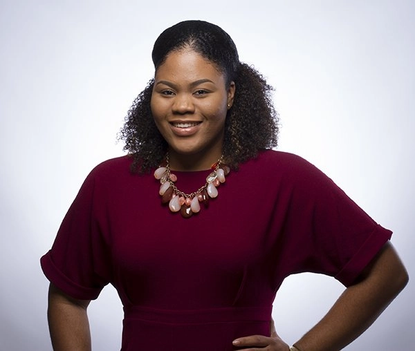 A studio portrait of a young African American woman with curly dark hair, wearing a solid cranberry-red dress and a chunky pink and coral beaded necklace. She is smiling confidently with one hand on her hip, set against a light gray background.