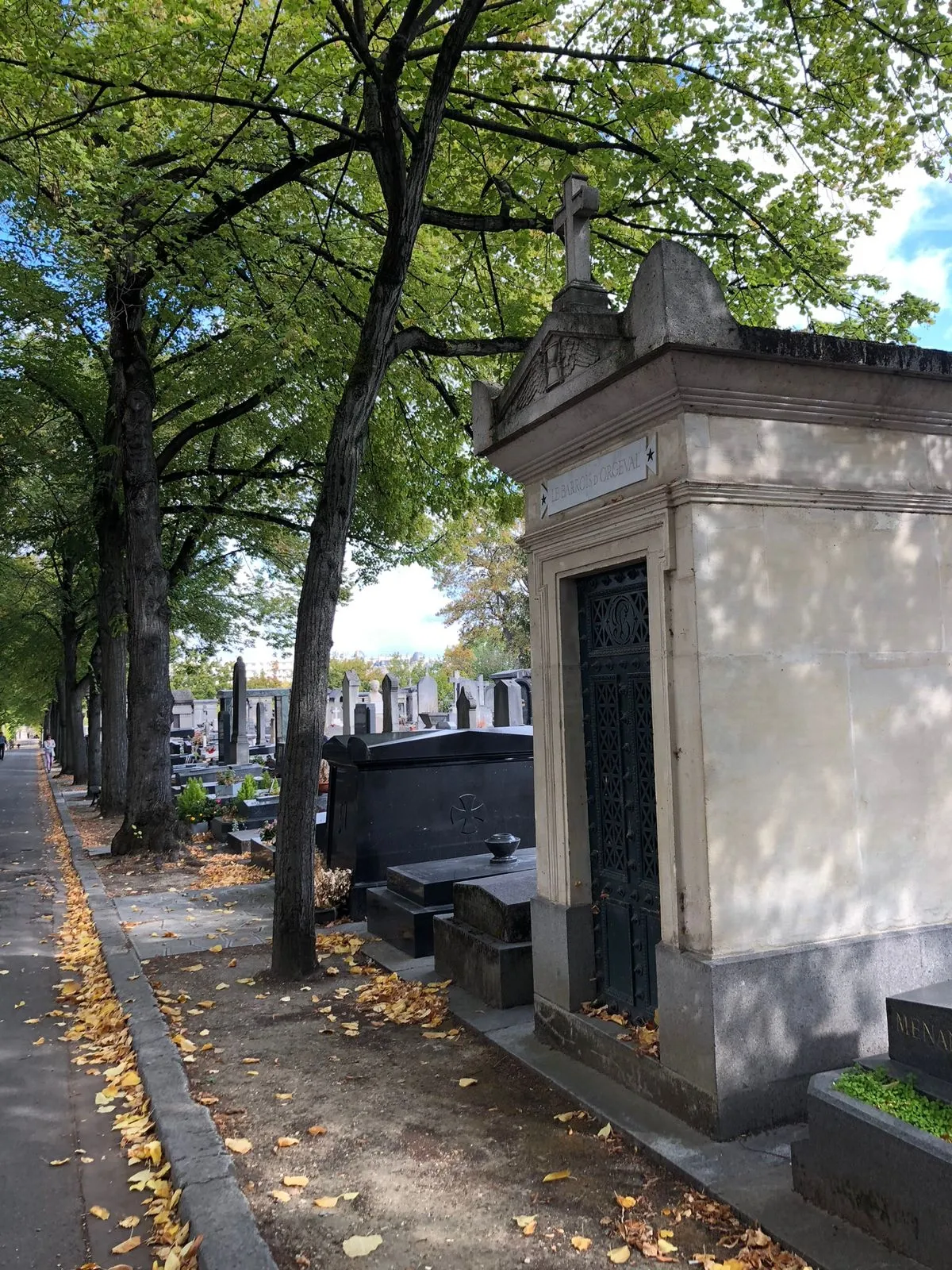 A photo taken in a large, leafy cemetery, likely in autumn, showing a long tree-lined path or alley on the left scattered with yellow leaves. On the right, a row of ornate stone tombs and mausoleums are visible. In the foreground, there is a prominent light-colored stone chapel-like mausoleum with a dark metal door and a stone cross on the roof.