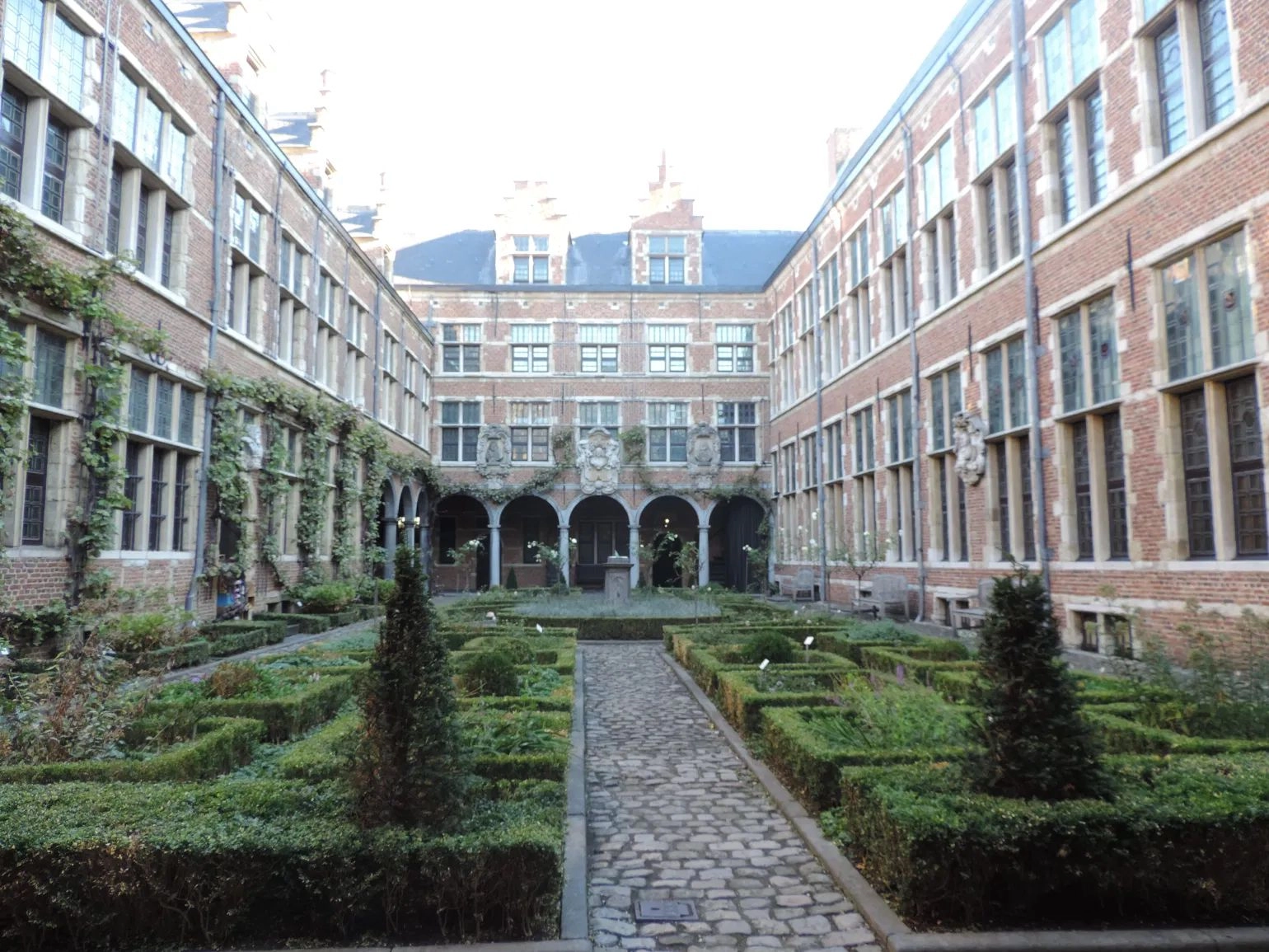 A tranquil view of a formal European courtyard garden surrounded by tall, three-story brick buildings with numerous multi-paned windows. The garden features a symmetrical design with a central cobblestone path flanked by manicured boxwood hedges and small topiaries. In the center-rear, an arched stone arcade leads to the building's inner structure.