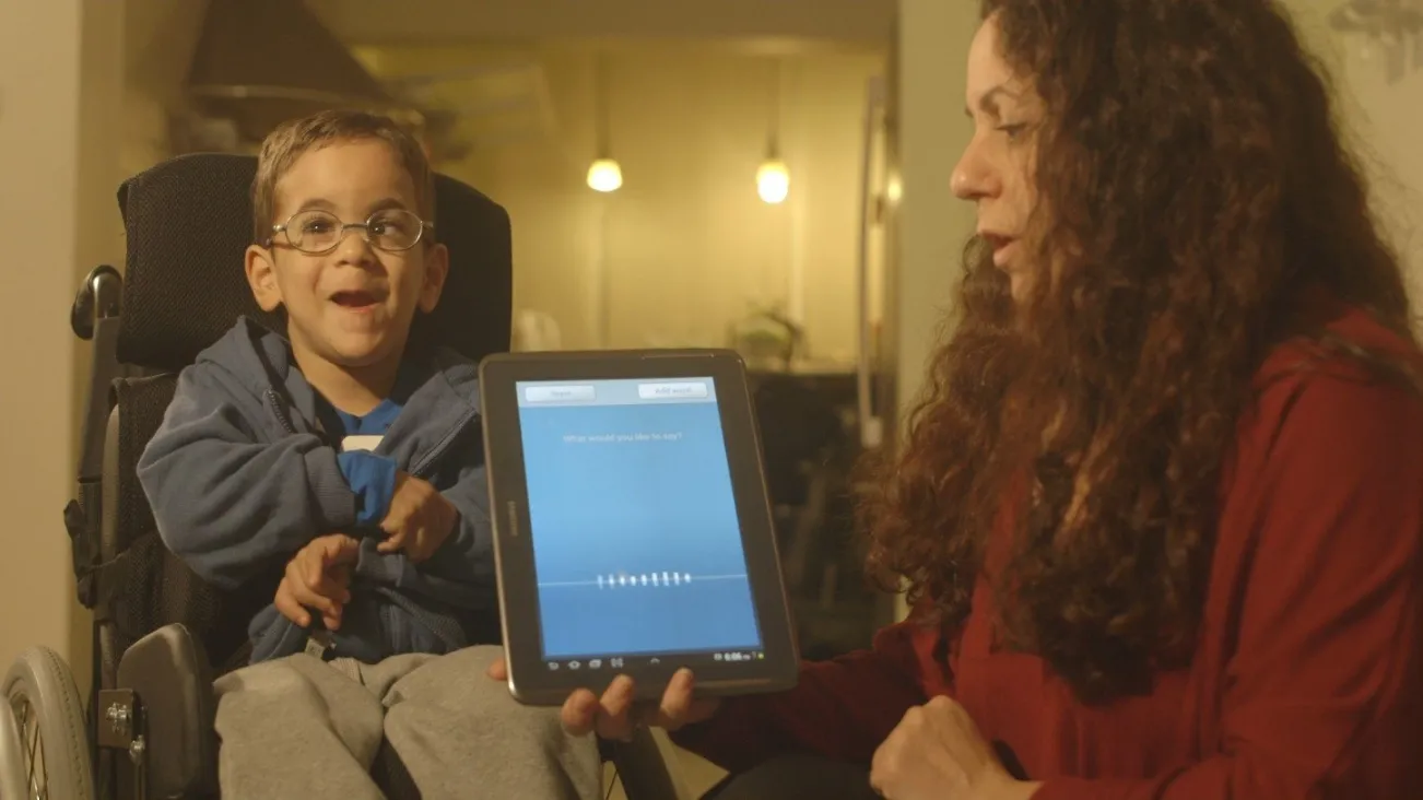 A warmly lit, medium shot of a woman with long, curly brown hair, wearing a red top, holding a tablet displaying an application screen. She is showing the screen to a young boy with glasses, who is seated in a wheelchair and smiling brightly. The boy is wearing a blue hooded sweatshirt. They appear to be indoors, possibly using an assistive communication technology.