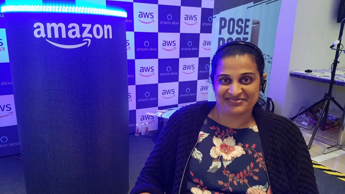 A professional portrait of a woman wearing a floral-patterned dress, a dark cardigan, and a headset, smiling at the camera. She is seated in front of a large, black column-shaped object displaying the "amazon" logo with a glowing blue light around the top edge, likely representing an Alexa device. The backdrop is a repeating pattern of "aws" and "amazon alexa" logos.