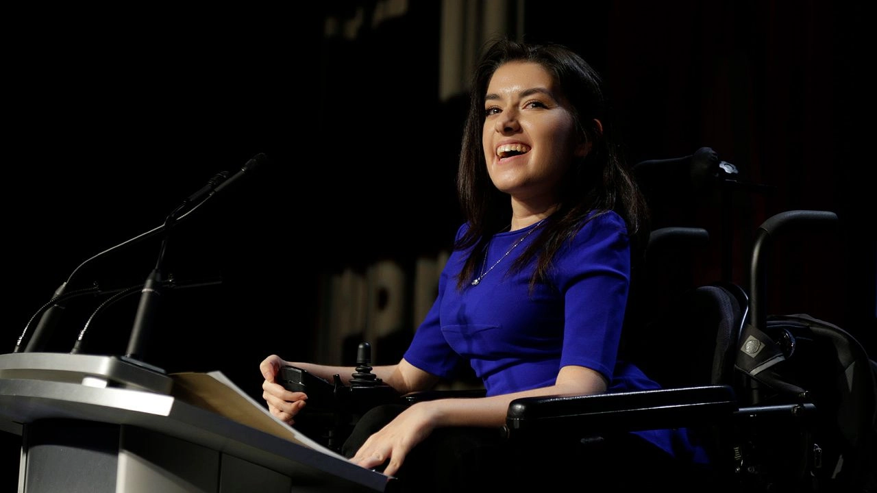 A young woman with dark hair, wearing a vibrant blue dress, is seated in a power wheelchair at a brightly lit podium, speaking into a microphone at a large event. The background is dark.